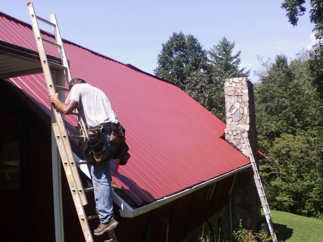 Roofer on a ladder beside a red metal roof and stone chimney — Soffit & Fascia Repair by Roofing Brothers in Calamus